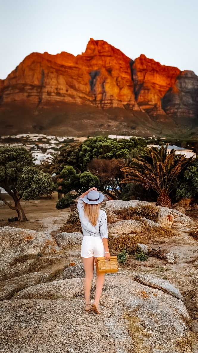 A woman in a sun hat looking out at the Twelve Apostles glowing orange at sunset above Camps Bay